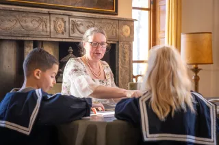 Twee kinderen in blauwe tenues zitten aan tafel en krijgen aanwijzingen van een volwassene
